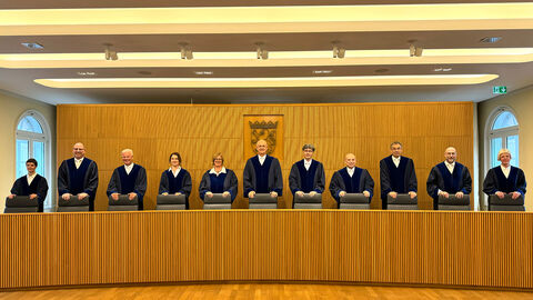 Die Mitglieder des Staatsgerichtshofs stehen nebeneinander hinter der Richterbank des Staatsgerichtshofs.